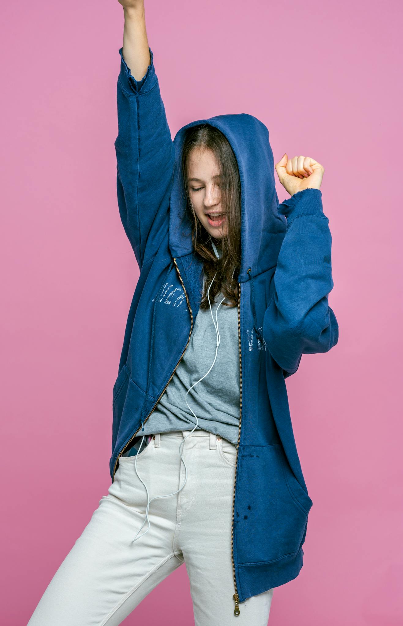 A smiling young woman dances in a blue hoodie with earphones against a pink background, embracing music and style.