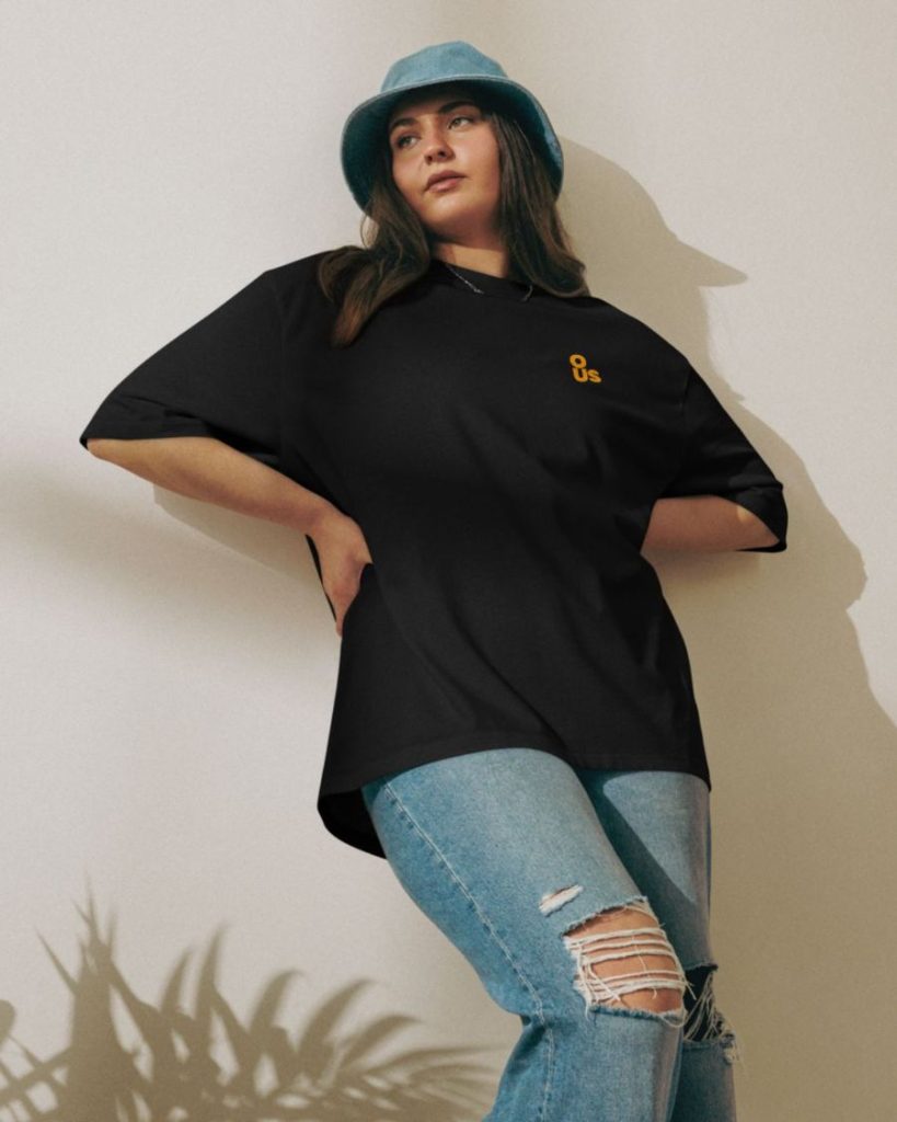Woman in a black oversized OUS logo tee and bucket hat leaning against a sunlit wall